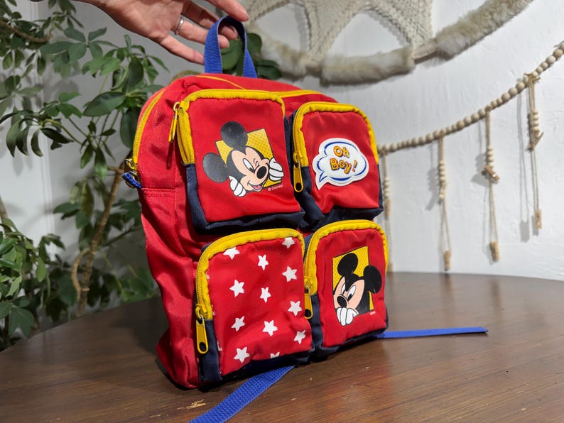 May include: Red backpack featuring Mickey Mouse and star graphics. The backpack has multiple zippered compartments with yellow trim and blue accents. One pocket has the text "Oh Boy!"