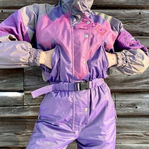 Peut inclure: Combinaison de ski vintage dans les tons de violet, rose et gris. La combinaison une pièce comprend une veste à capuche avec un motif géométrique et un pantalon assorti. Elle a une ceinture avec une boucle noire et des poignets élastiques.