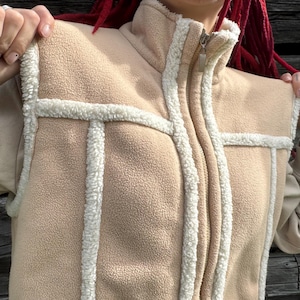 Op de afbeelding: Een beige fleecevest met een hoge kraag en een ritssluiting over de gehele lengte. Het kledingstuk is afgewerkt met witte sherpa-randen langs de naden, armsgaten en kraag, wat zorgt voor een gestructureerde uitstraling en zachte textuur.