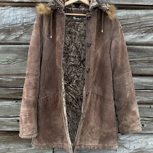 May include: A brown suede coat with a fur-lined hood and interior. The coat has a button closure, a zipper, and two front pockets. The coat is hanging on a wooden hanger against a wooden background.