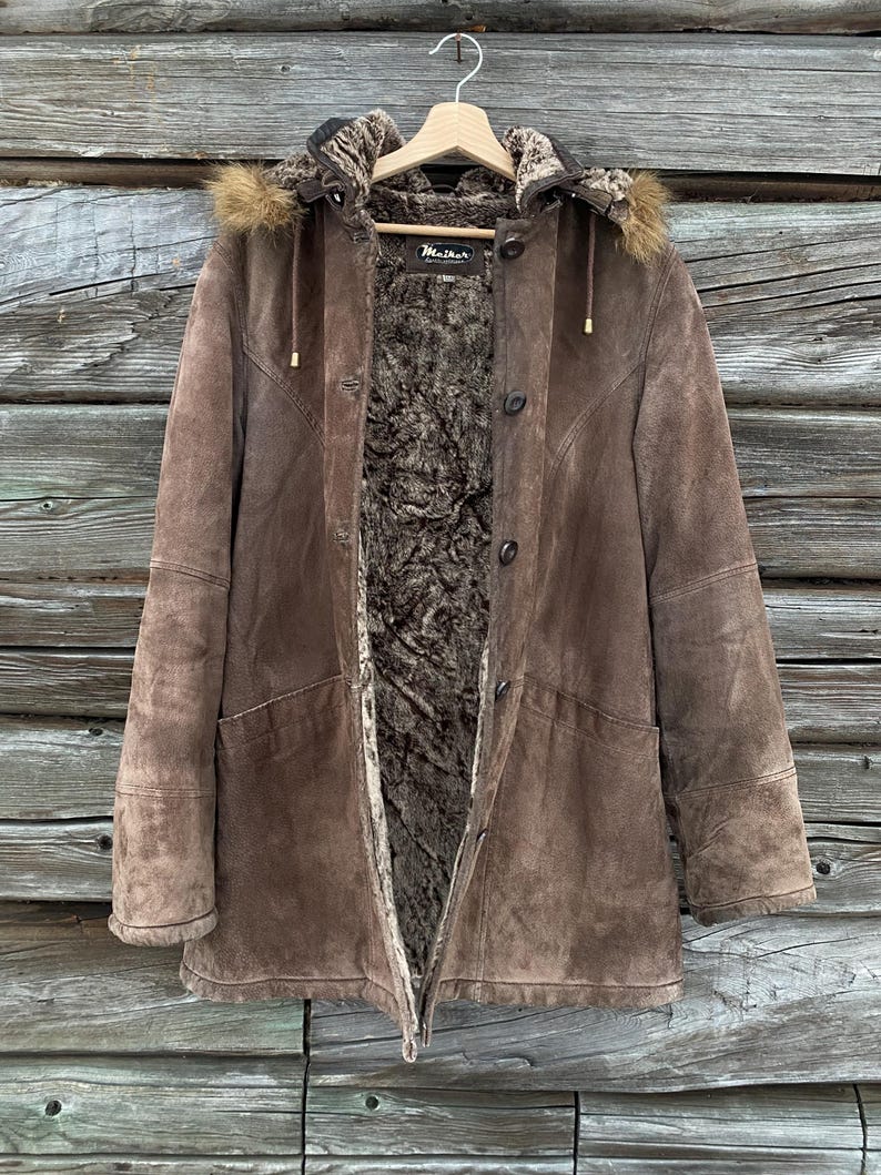 May include: A brown suede coat with a fur-lined hood and interior. The coat has a button closure, a zipper, and two front pockets. The coat is hanging on a wooden hanger against a wooden background.