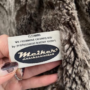 May include: Close-up of a plush, textured fabric in shades of brown and gray. A white tag with the word "CLEANING" and cleaning instructions is visible. The tag also features the brand name "Meiker".