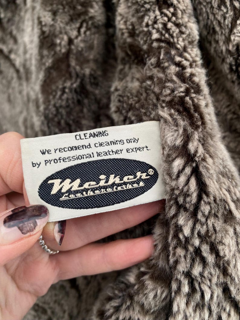 May include: Close-up of a plush, textured fabric in shades of brown and gray. A white tag with the word "CLEANING" and cleaning instructions is visible. The tag also features the brand name "Meiker".