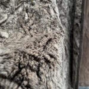 May include: Close-up of a brown and gray faux fur jacket. The texture is dense and shaggy, with varying shades of brown and gray. The jacket has a zipper and a brown leather-like trim.