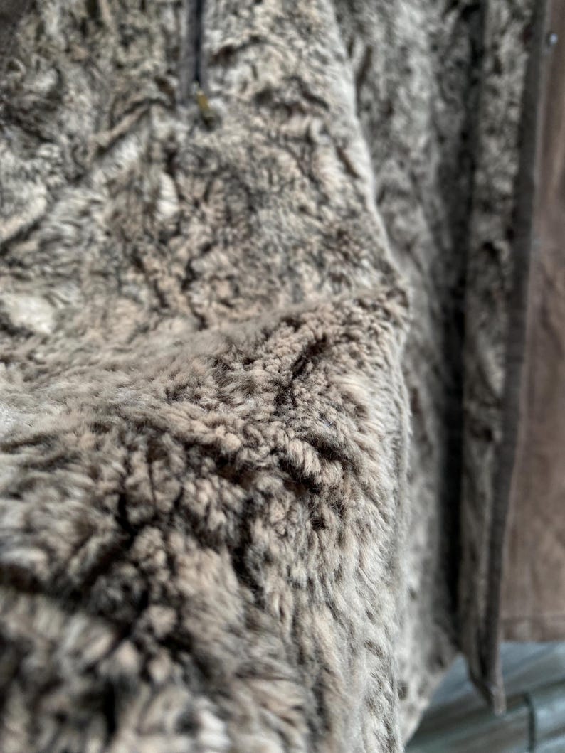 May include: Close-up of a brown and gray faux fur jacket. The texture is dense and shaggy, with varying shades of brown and gray. The jacket has a zipper and a brown leather-like trim.