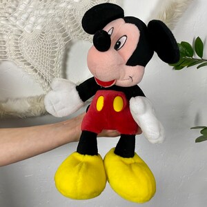 May include: A plush Mickey Mouse toy with black ears, a red shirt with yellow buttons, and yellow shoes. The toy is being held by a person's hand.