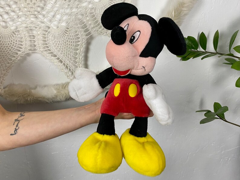 May include: A plush Mickey Mouse toy with black ears, a red shirt with yellow buttons, and yellow shoes. The toy is being held by a person's hand.