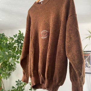 May include: A brown, oversized sweater with a crew neck and long sleeves. The sweater features a textured knit and a circular emblem on the chest that reads "ENGLISH STONE". The garment is hanging on a wooden hanger.