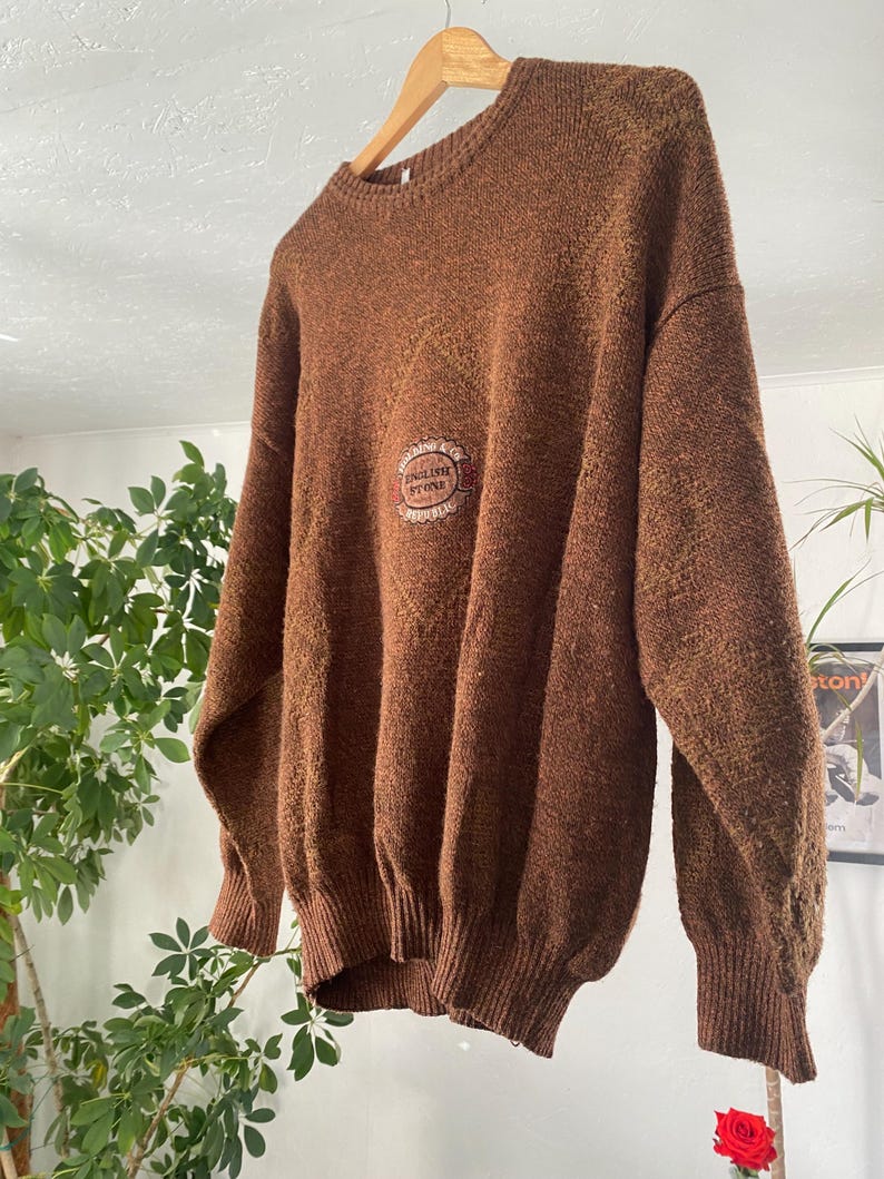 May include: A brown, oversized sweater with a crew neck and long sleeves. The sweater features a textured knit and a circular emblem on the chest that reads "ENGLISH STONE". The garment is hanging on a wooden hanger.