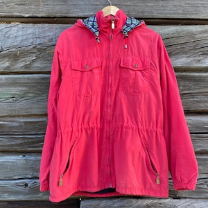 May include: A bright red jacket with a hood and a full-length zipper. The jacket has two chest pockets and two lower pockets, all with zippers. The hood lining features a blue and green plaid pattern. The jacket is hanging on a wooden hanger.