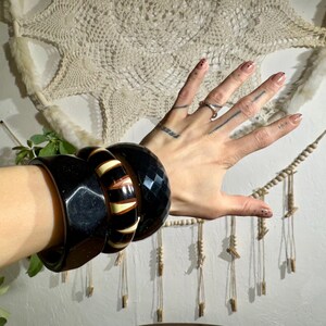 Op de afbeelding: Een verzameling zwarte en bruine armbanden om een pols. De armbanden hebben geometrische en gestreepte ontwerpen. De achtergrond omvat een crèmekleurige wandhanger en groen.