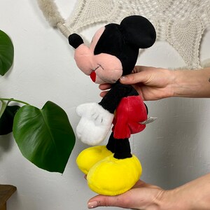 May include: A plush Mickey Mouse toy with black fur, red shorts, white gloves, and yellow shoes. The toy is standing on its feet and has a friendly expression.