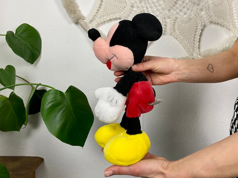 May include: A plush Mickey Mouse toy with black fur, red shorts, white gloves, and yellow shoes. The toy is standing on its feet and has a friendly expression.