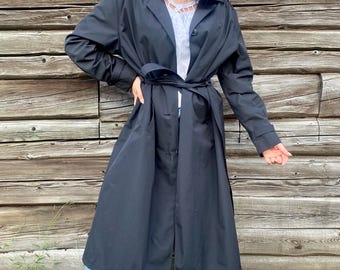 Puede incluir: Una gabardina negra con cinturón y cierre de botones. La gabardina está hecha de una tela ligera y tiene un diseño clásico.