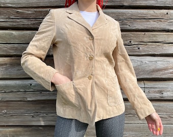 Könnte beinhalten: Eine hellbraune Wildlederjacke mit einem Reverskragen, drei Knöpfen und zwei Pattentaschen. Die Jacke hat eine leicht verkürzte Länge und eine lockere Passform.