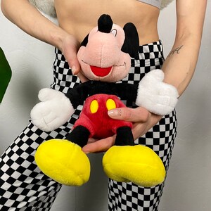 May include: A plush Mickey Mouse toy with a red and yellow outfit. The toy is being held by a person wearing black and white checkered pants.