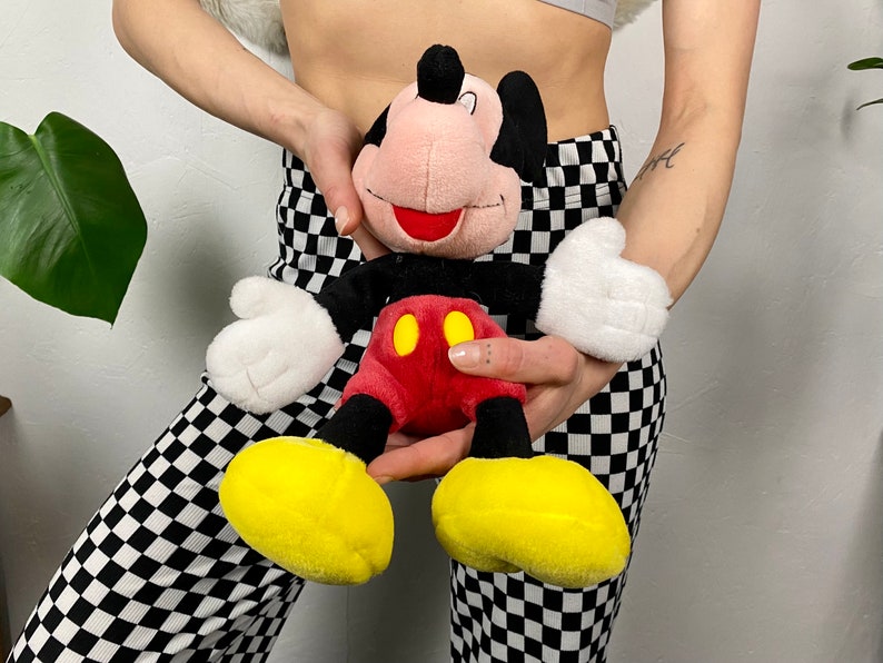 May include: A plush Mickey Mouse toy with a red and yellow outfit. The toy is being held by a person wearing black and white checkered pants.