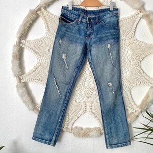 May include: Distressed blue denim jeans with a light wash and multiple rips and tears throughout. The jeans have a classic five-pocket style and a straight leg.