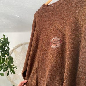 May include: Brown knit sweater with a crew neck and long sleeves. The sweater features a small embroidered patch with the words "ENGLISH STONE" in a circular design. The sweater is hanging on a wooden hanger.