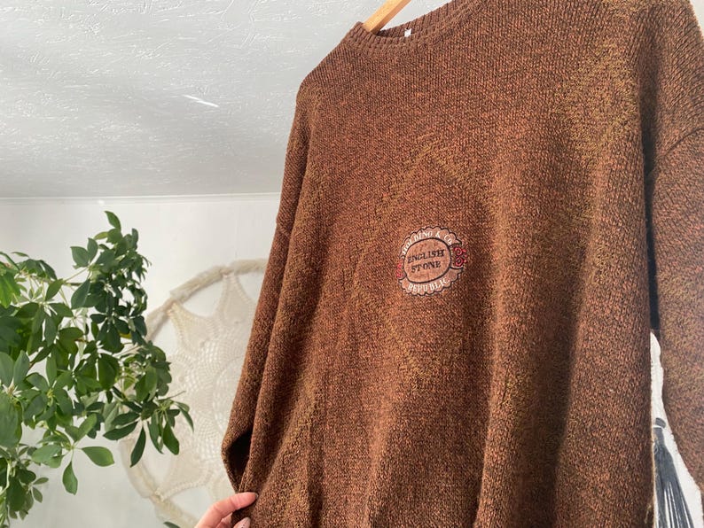 May include: Brown knit sweater with a crew neck and long sleeves. The sweater features a small embroidered patch with the words "ENGLISH STONE" in a circular design. The sweater is hanging on a wooden hanger.