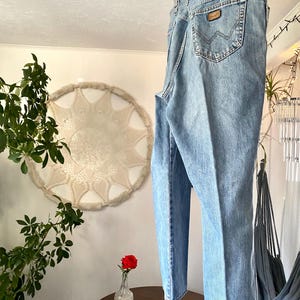 May include: Light blue denim jeans hanging on a wooden hanger. The jeans have a classic five-pocket design, a brown leather patch on the back pocket, and a straight leg fit. The jeans are made of a medium-wash denim.