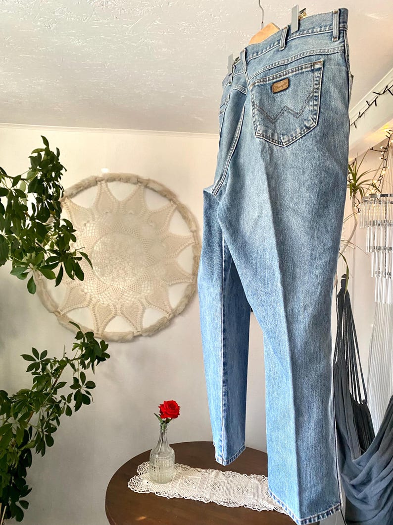 May include: Light blue denim jeans hanging on a wooden hanger. The jeans have a classic five-pocket design, a brown leather patch on the back pocket, and a straight leg fit. The jeans are made of a medium-wash denim.