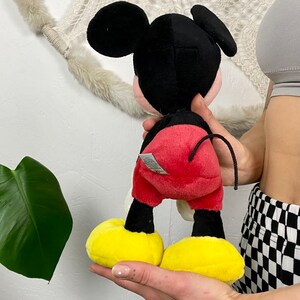 May include: A plush Mickey Mouse toy with black ears, a black head, red shorts, yellow shoes, and a black tail.