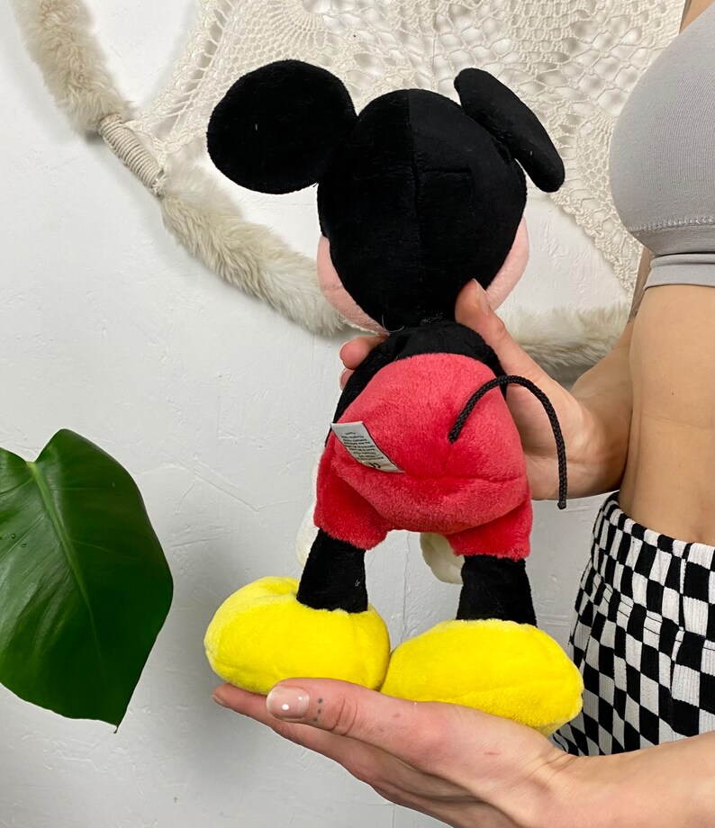 May include: A plush Mickey Mouse toy with black ears, a black head, red shorts, yellow shoes, and a black tail.