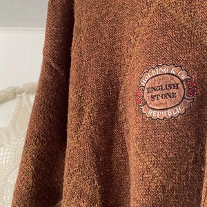 May include: A brown sweater with a textured knit. The sweater features a circular patch with the words "HOLDING & Co. ENGLISH STONE REPUBLIC" in a decorative design. The sweater has a classic, casual style.