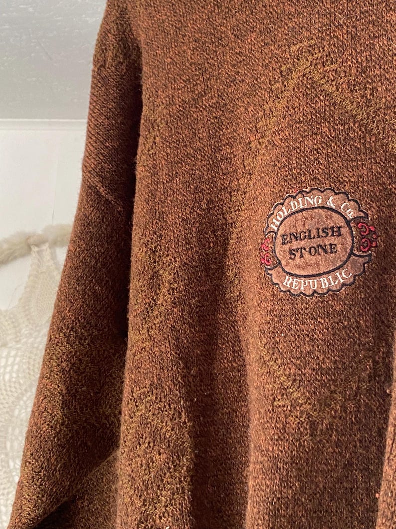 May include: A brown sweater with a textured knit. The sweater features a circular patch with the words "HOLDING & Co. ENGLISH STONE REPUBLIC" in a decorative design. The sweater has a classic, casual style.