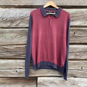 Può includere: Polo a maniche lunghe bordeaux e blu navy. Il corpo della camicia presenta un motivo strutturato, mentre le maniche e il colletto sono blu navy. La camicia è appesa a una gruccia di legno su uno sfondo di legno invecchiato.