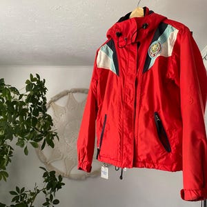 May include: A vibrant red hooded jacket with black and white detailing. The jacket features a zip closure, two front pockets, and a logo patch on the upper left chest. It is suspended from a wooden hanger.
