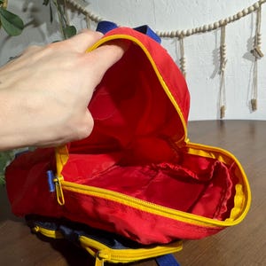 May include: A small, red backpack with yellow trim and a blue zipper. The interior is red. The backpack has blue straps and is open, revealing the inside. The backpack is on a wooden surface.