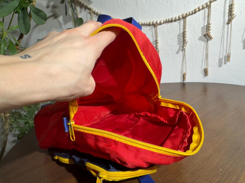 May include: A small, red backpack with yellow trim and a blue zipper. The interior is red. The backpack has blue straps and is open, revealing the inside. The backpack is on a wooden surface.