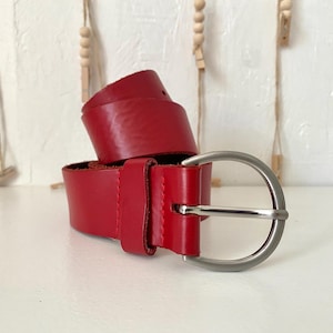 May include: A bright red belt featuring a smooth, solid-colored strap and a polished silver-toned metal buckle. The belt is coiled, showing a single prong and a matching red leather loop to secure the end of the strap once it is fastened.