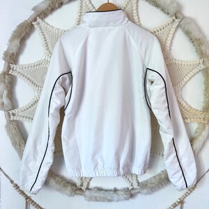 May include: A white jacket with a high collar and black trim. The jacket has long sleeves and a drawstring hem. It is hanging on a wooden hanger against a decorative backdrop.
