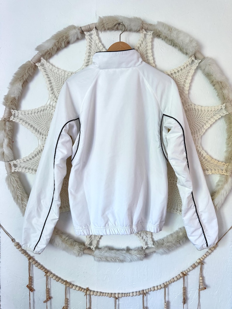 May include: A white jacket with a high collar and black trim. The jacket has long sleeves and a drawstring hem. It is hanging on a wooden hanger against a decorative backdrop.