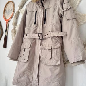 May include: A beige long-length coat featuring a faux fur collar, a waist belt with a buckle, multiple utility pockets with flaps, and vertical zippered chest pockets. The coat has long sleeves with ribbed cuffs and a button-down front closure.