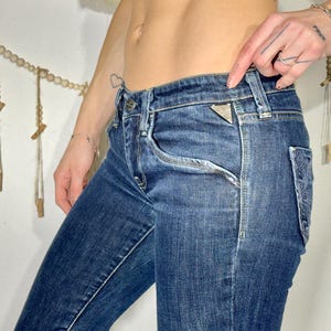 May include: Dark wash denim jeans with a classic five-pocket design. The jeans feature a triangle logo on the front pocket and decorative stitching on the back pocket. The jeans are a slim fit.