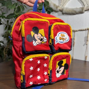 May include: Red Mickey Mouse backpack with multiple zippered pockets. Features Mickey Mouse graphics and white star patterns. Includes blue straps and yellow zippers. The backpack has a blue handle and the text "Oh Boy!"