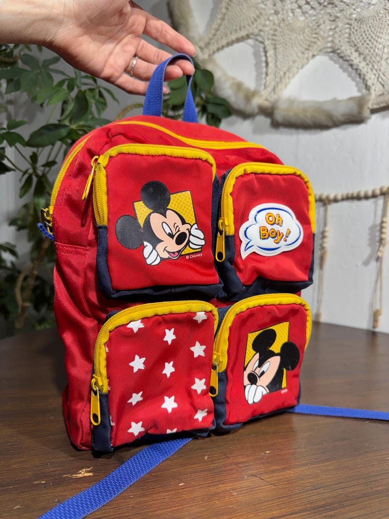 May include: Red Mickey Mouse backpack with multiple zippered pockets. Features Mickey Mouse graphics and white star patterns. Includes blue straps and yellow zippers. The backpack has a blue handle and the text "Oh Boy!"