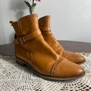 K&ouml;nnte beinhalten: Ein Paar hellbraune Leder-Ankle Boots mit Ziern&auml;hten und Schnallenverschl&uuml;ssen. Die Stiefel haben einen niedrigen Absatz und eine runde Spitze. Die Stiefel stehen auf einer wei&szlig;en Spitzentischdecke. Die Stiefel sind ca. 15 cm hoch.