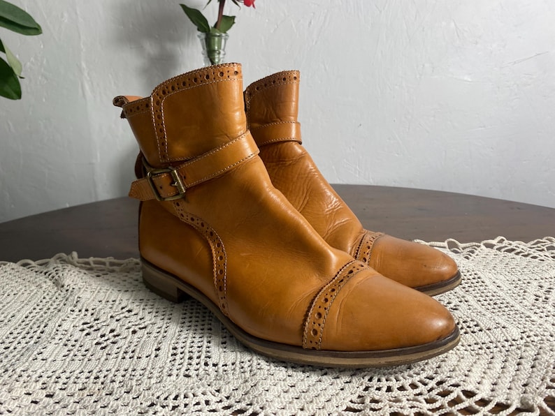 K&ouml;nnte beinhalten: Ein Paar hellbraune Leder-Ankle Boots mit Ziern&auml;hten und Schnallenverschl&uuml;ssen. Die Stiefel haben einen niedrigen Absatz und eine runde Spitze. Die Stiefel stehen auf einer wei&szlig;en Spitzentischdecke. Die Stiefel sind ca. 15 cm hoch.