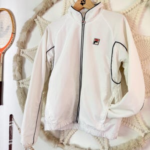 May include: White zip-up jacket with black trim and a small logo on the chest. The jacket has a high collar and long sleeves. The jacket is hanging on a wooden hanger.