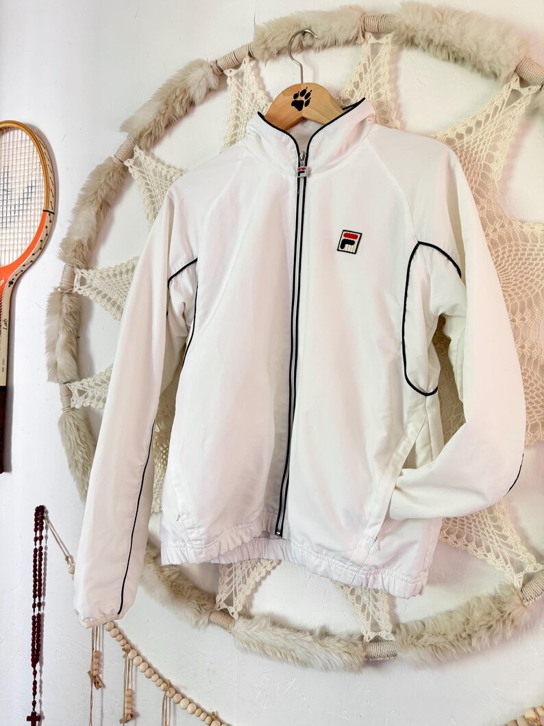 May include: White zip-up jacket with black trim and a small logo on the chest. The jacket has a high collar and long sleeves. The jacket is hanging on a wooden hanger.
