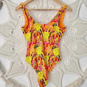 May include: A one-piece swimsuit featuring a vibrant pattern of yellow SpongeBob SquarePants characters on a pink, orange, and yellow background. The swimsuit has a V-neckline and is hanging on a wooden hanger.