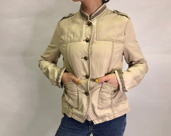 May include: A beige military-style jacket with a stand-up collar, button closure, and decorative trim on the sleeves. The jacket has two front pockets and a fitted silhouette.