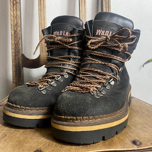 Pode incluir: Um par de botas de caminhada de camurça preta com atacadores castanhos e uma sola grossa, castanho-claro e preta. O texto "WILD LAND" em rosa está na língua. As botas estão numa cadeira de madeira.