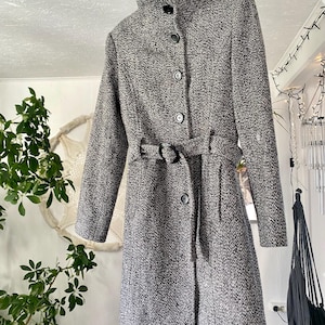 Può includere: Un cappotto in tweed grigio e bianco con collo alto e cintura in vita. Il cappotto ha una chiusura a bottoni sul davanti e maniche lunghe. Il cappotto è appeso a un'appendiabiti.