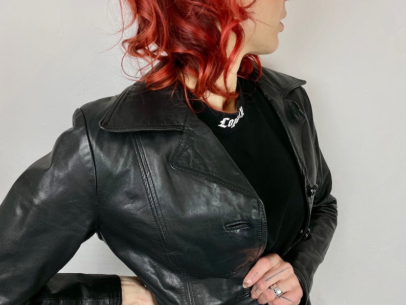 May include: A black leather jacket with a button closure and a collar. The jacket is worn by a person with red hair. The person is wearing a black t-shirt with the word "Loyal" written on it.